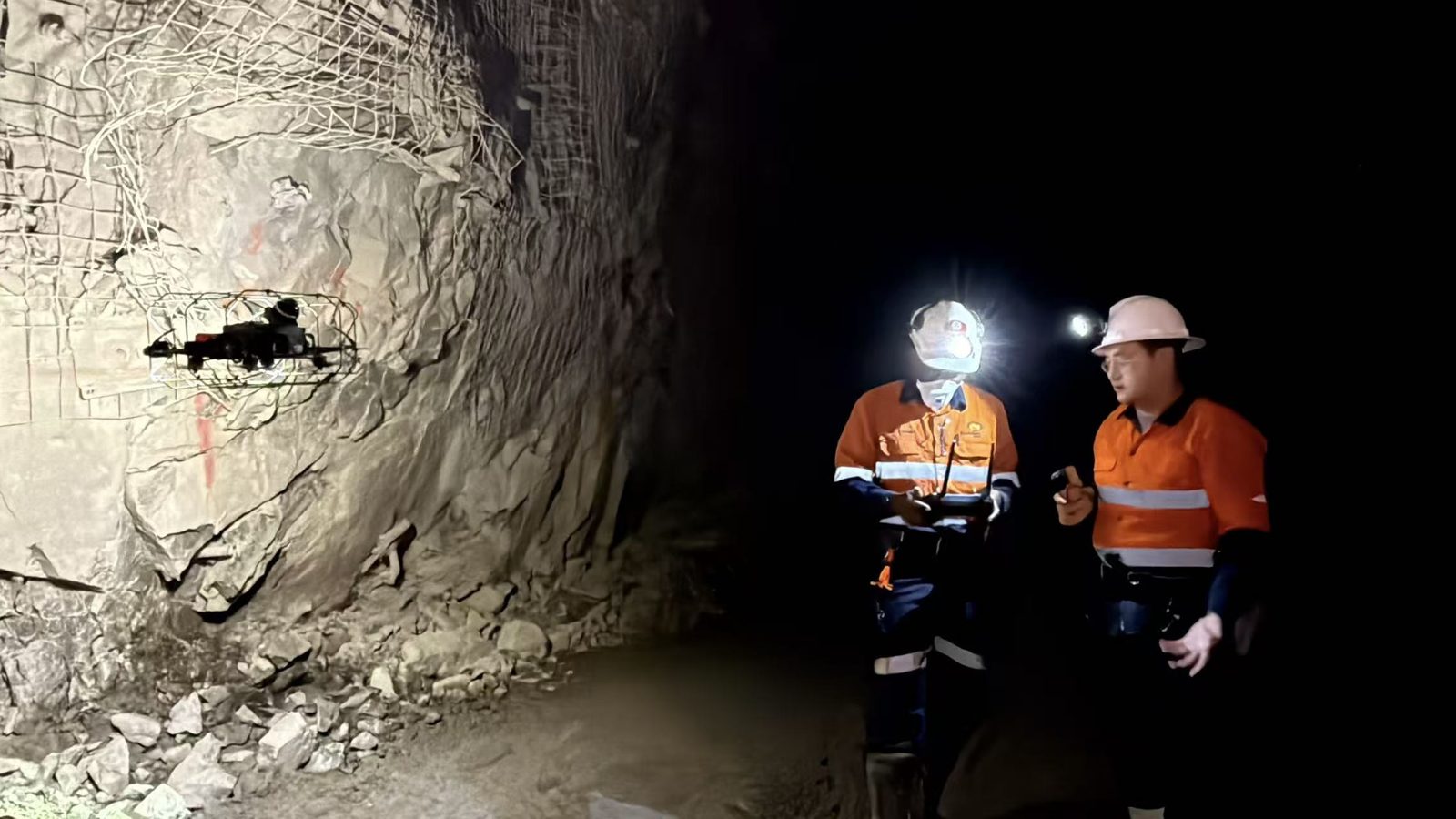 Kookaburra drone hovering beside a mesh-covered rock wall underground, with two crew in high-visibility gear watching